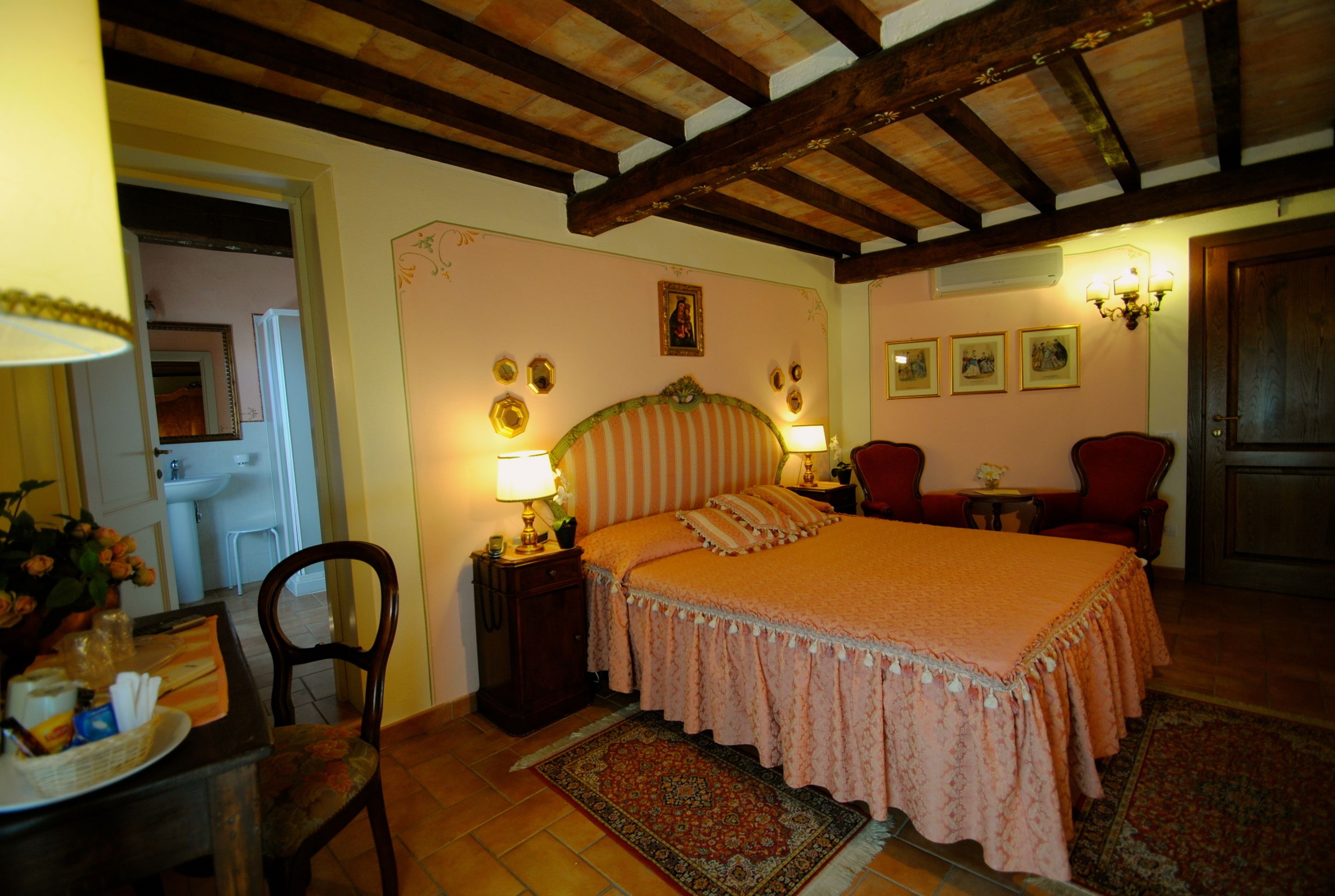 A bedroom with a large bed, wooden beams on the ceiling, a desk with a chair, and a small table with a lamp.