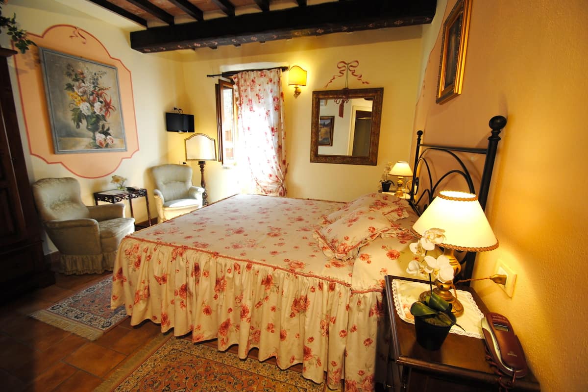 A bedroom with a floral bedspread, a bedside lamp, a chair, a small table, and a mirror on the wall.