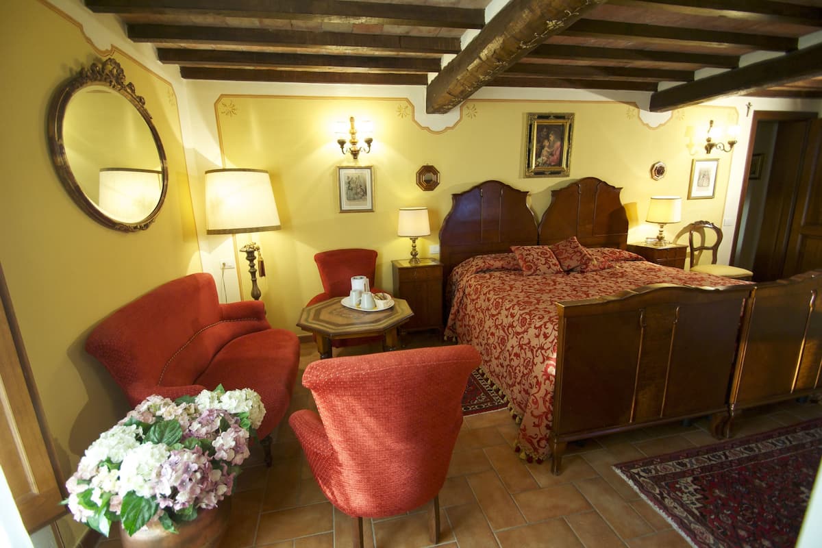 A traditional bedroom with a wooden bed, a red armchair, a small table with a lamp, and a mirror on the wall.