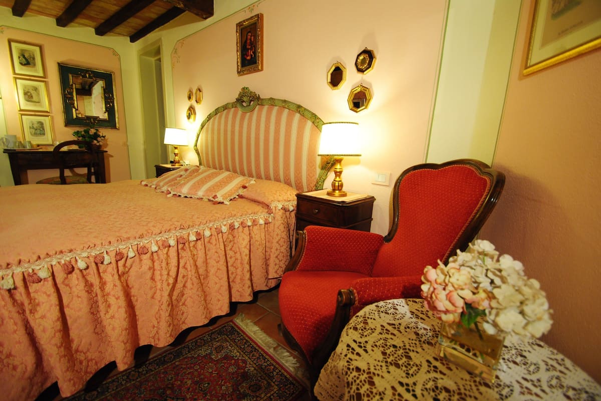 A bedroom with a bed, a red armchair, a nightstand with a lamp, a small table with a vase of flowers, and a framed picture on the wall.