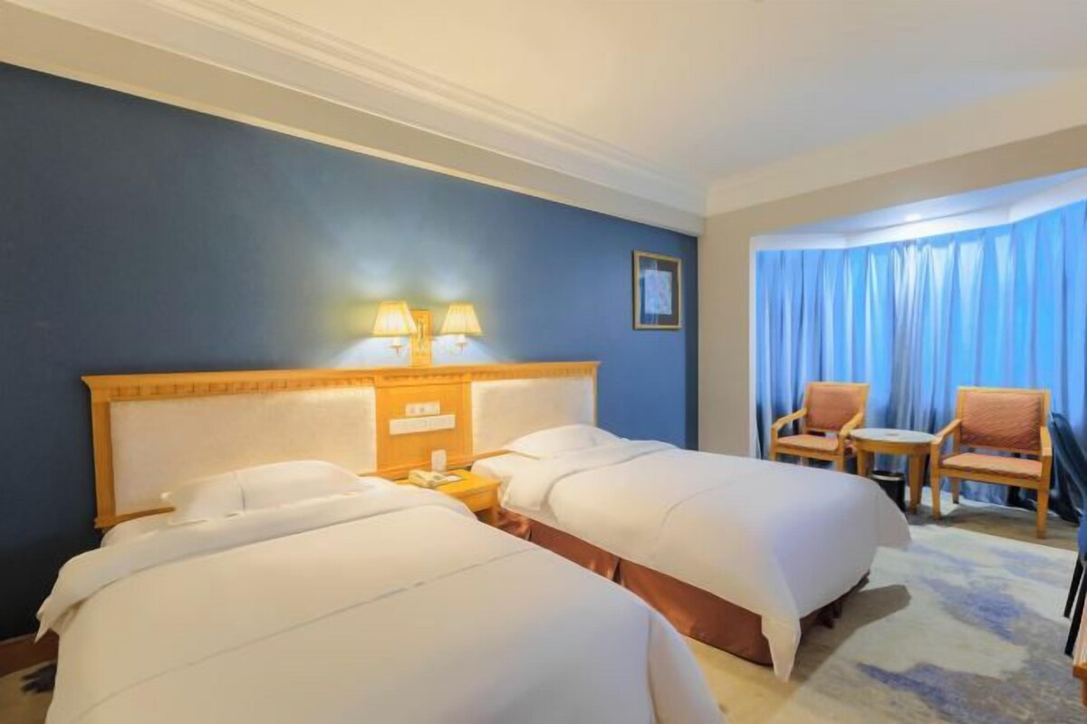 Habitación de hotel con dos camas, una mesita de noche con lámparas, una silla y vistas al exterior a través de cortinas.
