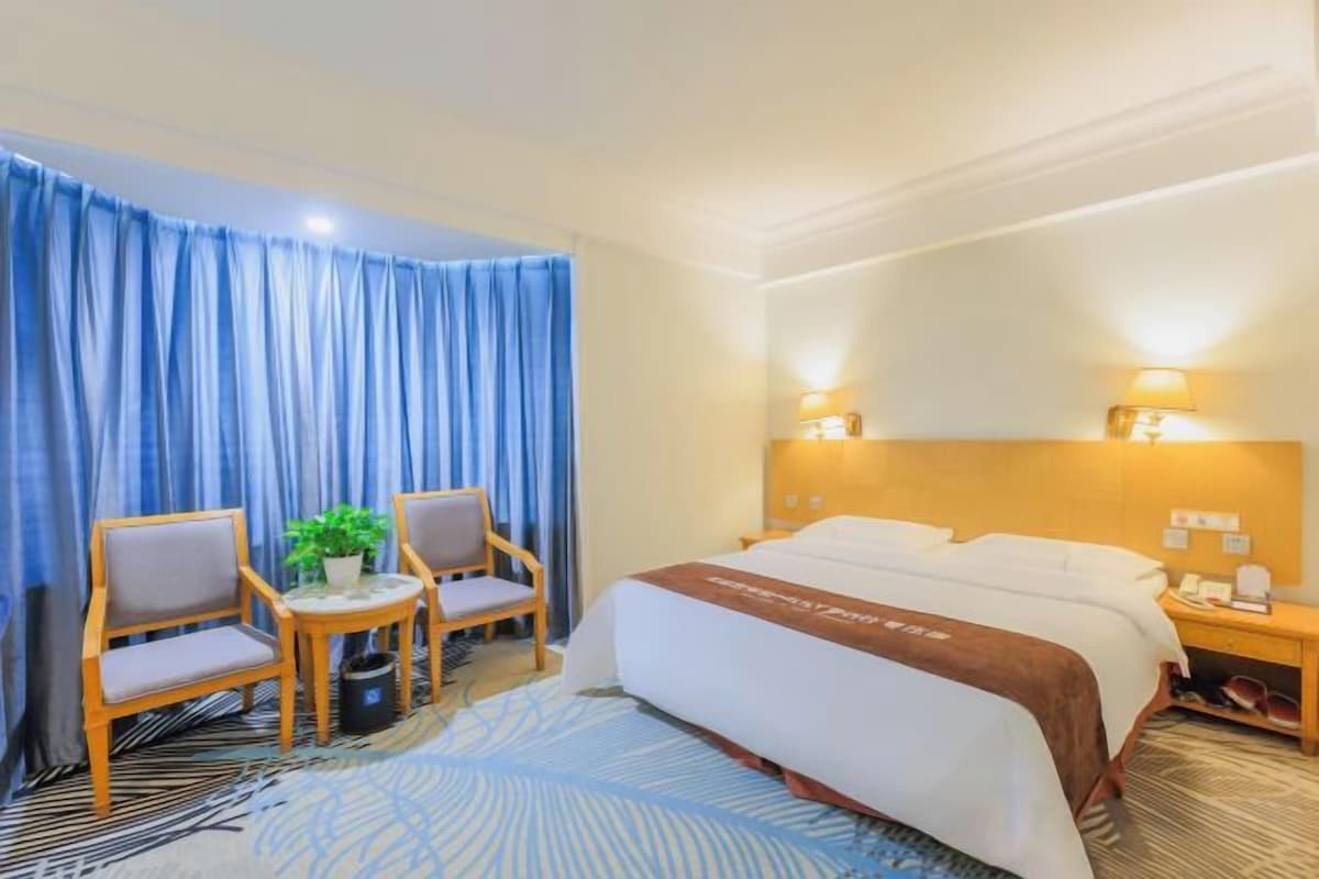A hotel room with a bed, two chairs, a small table with a plant, and a nightstand with a lamp.