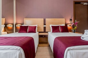 A hotel room with two beds, each with a wooden headboard, white bedding, and red throw pillows. There are bedside tables with lamps, a small potted plant, and a stack of towels.