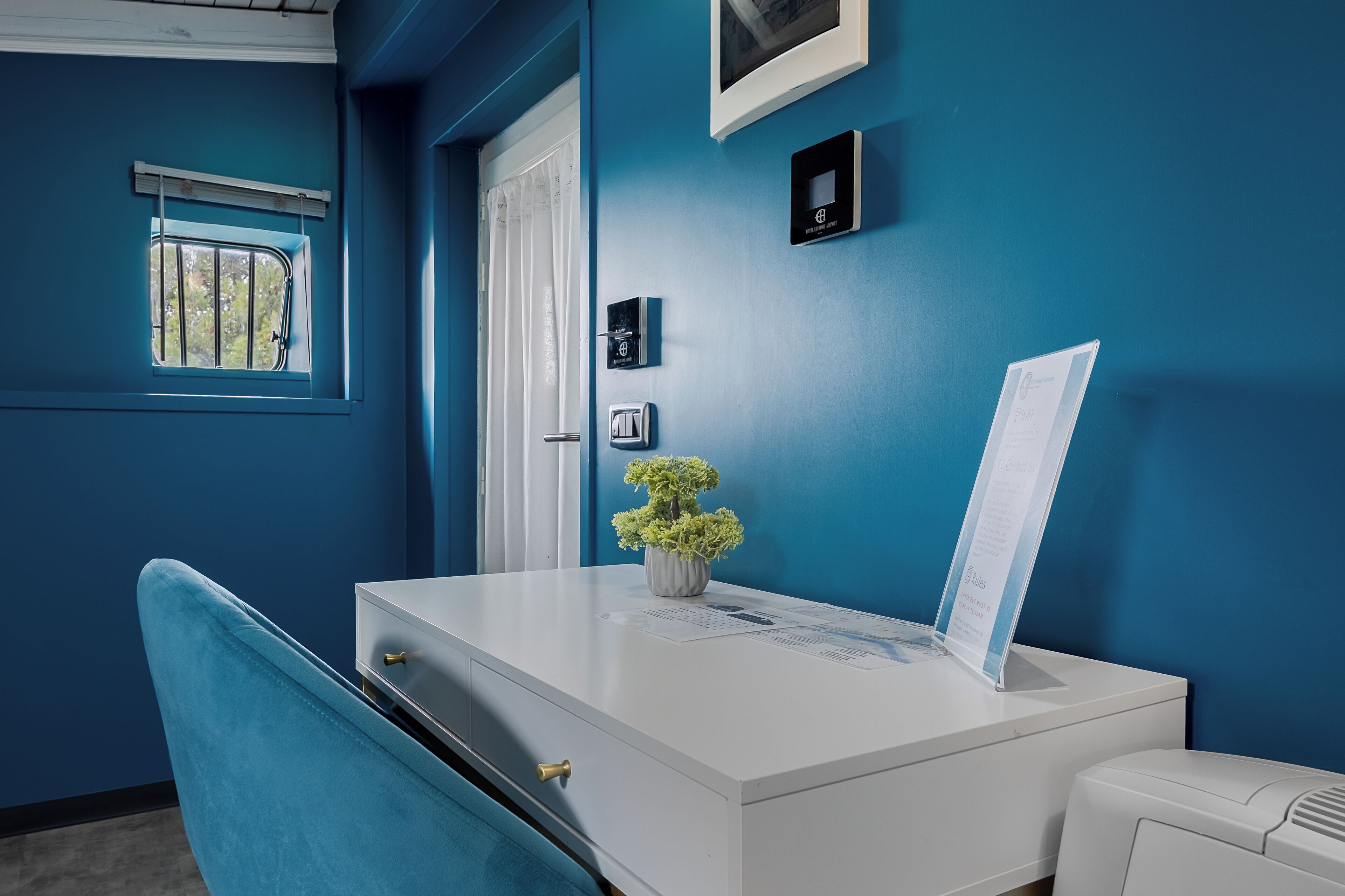 Ein Zimmer mit weißem Schreibtisch, blauem Stuhl, kleinem Fenster mit Gitterstäben, einer Tür und einem Thermostat an der Wand.