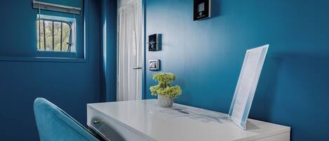 Ein Zimmer mit weißem Schreibtisch, blauem Stuhl, kleinem Fenster mit Gitterstäben, einer Tür und einem Thermostat an der Wand.