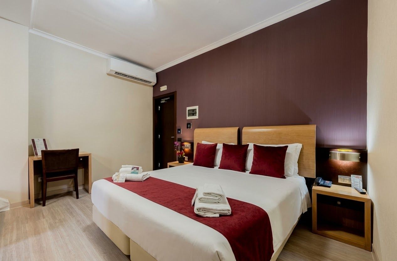 A hotel room with a bed, bedside table, desk, chair, and an air conditioning unit.