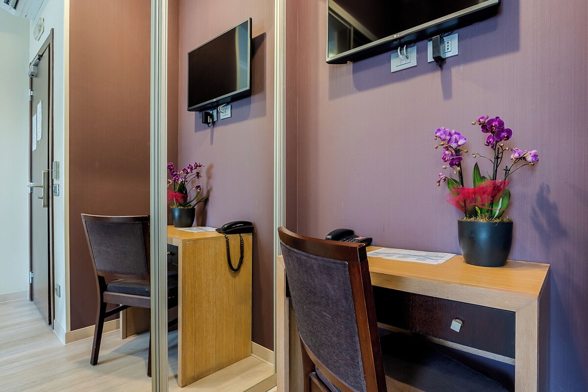 A hotel room with a wooden desk, a chair, a television mounted on the wall, and a potted orchid plant.
