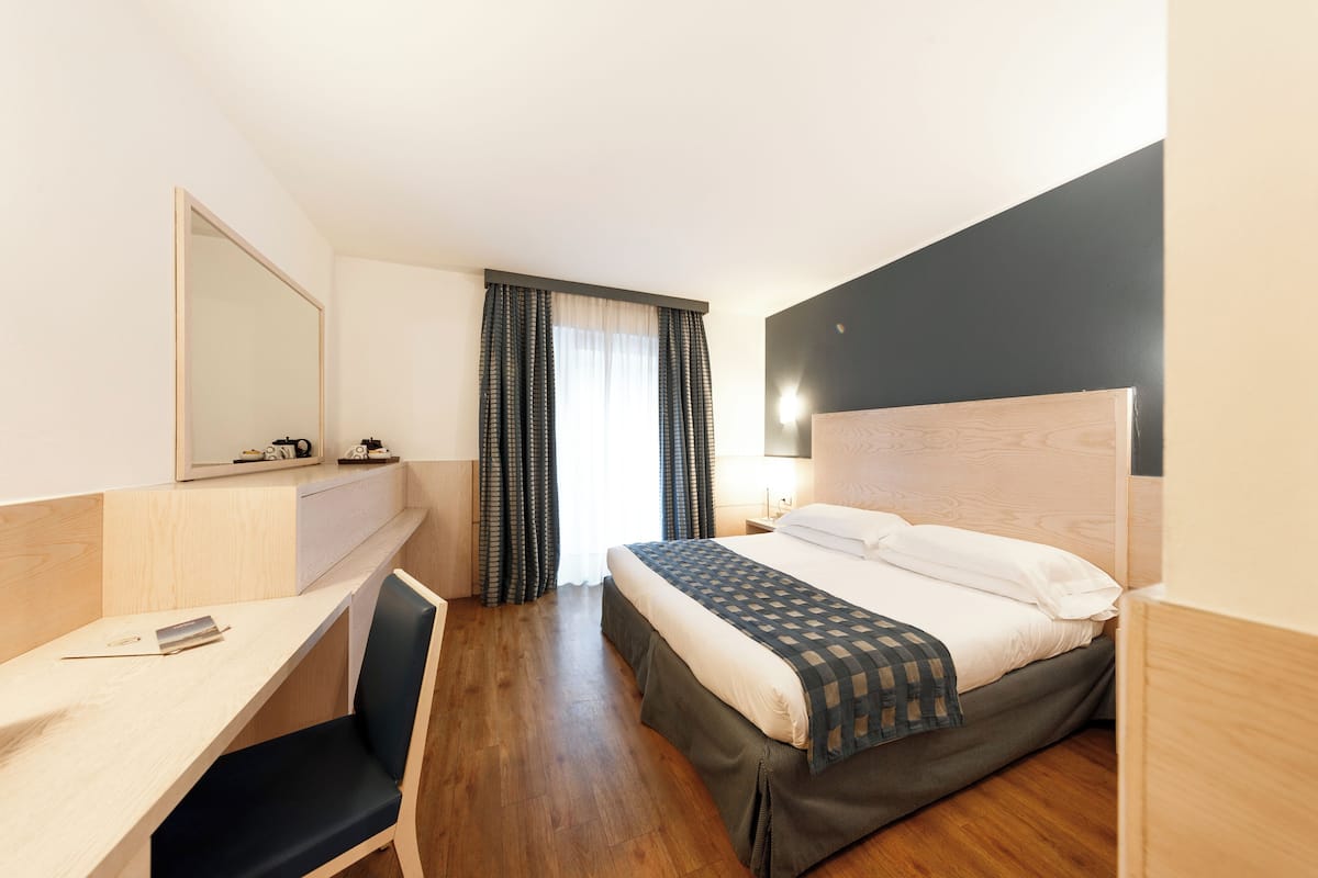 A hotel room with a bed, a desk, a chair, a mirror, and a window with curtains.