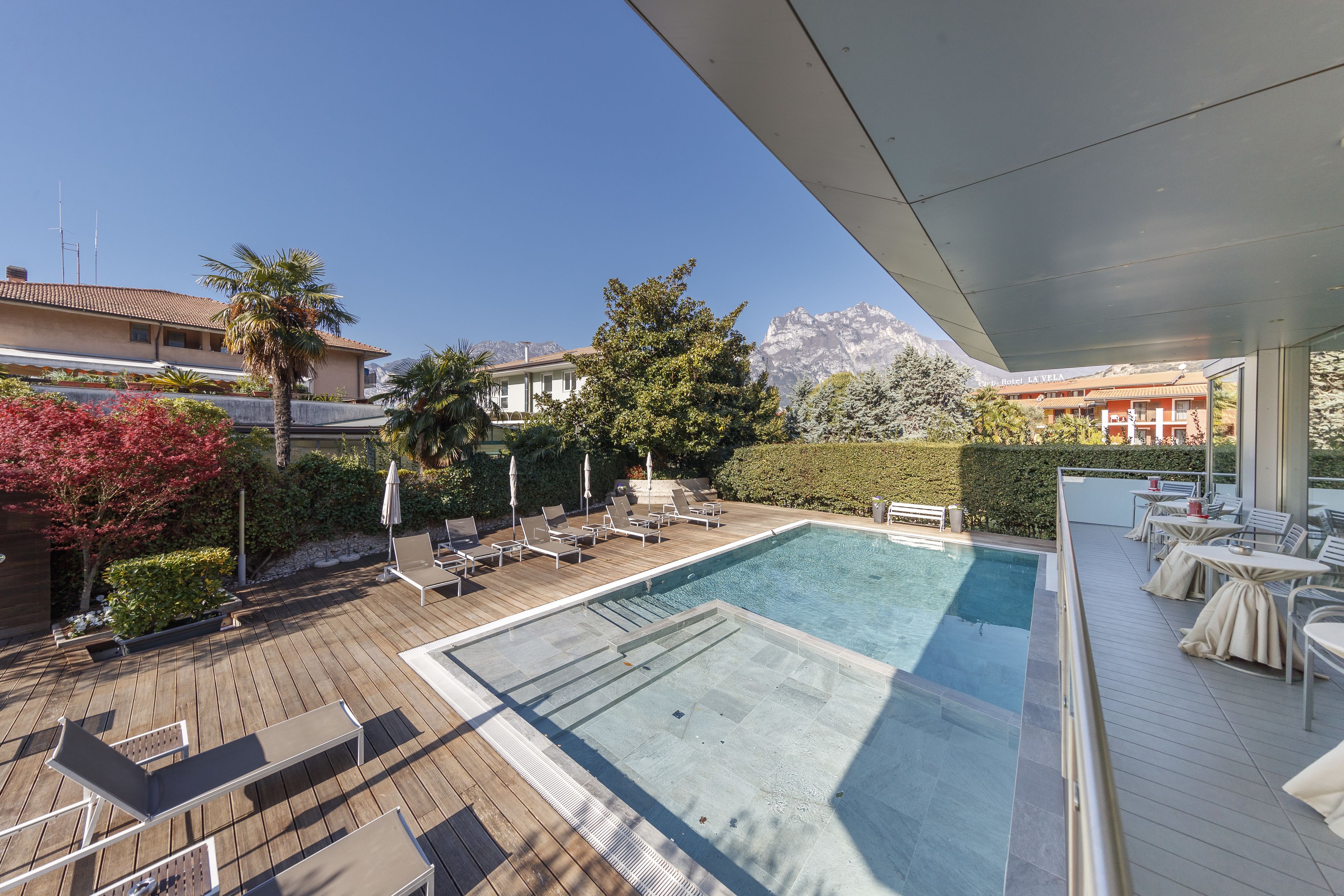 A rooftop pool area with lounge chairs and tables, surrounded by greenery and a view of mountains.