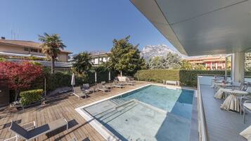 A rooftop pool area with lounge chairs and tables, surrounded by greenery and a view of mountains.