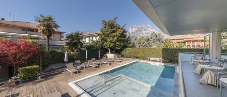 A rooftop pool area with lounge chairs and tables, surrounded by greenery and a view of mountains.