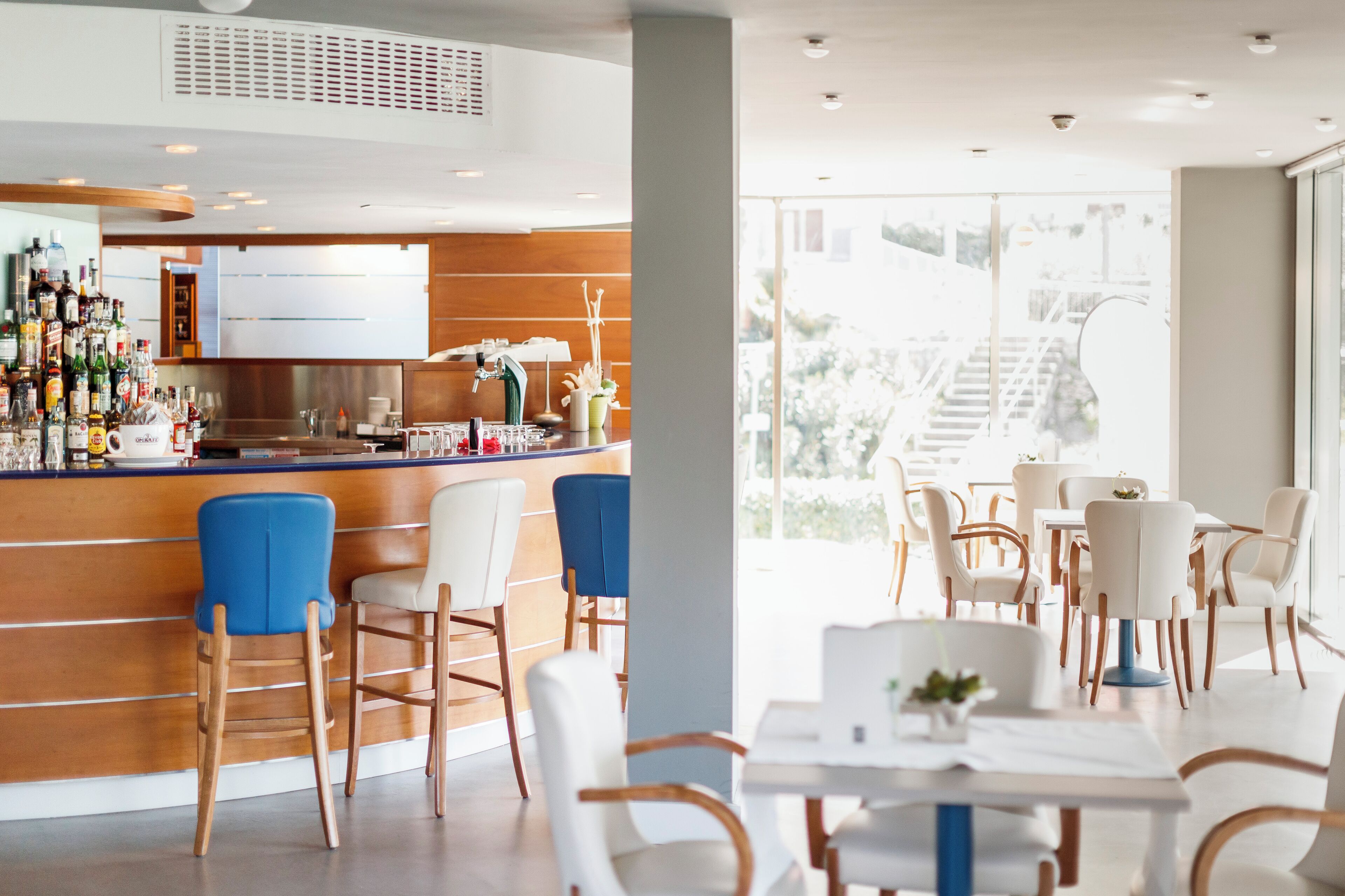 A modern bar area with wooden bar counter and blue chairs, a dining area with white tables and chairs, and large windows offering a view of the outdoors.
