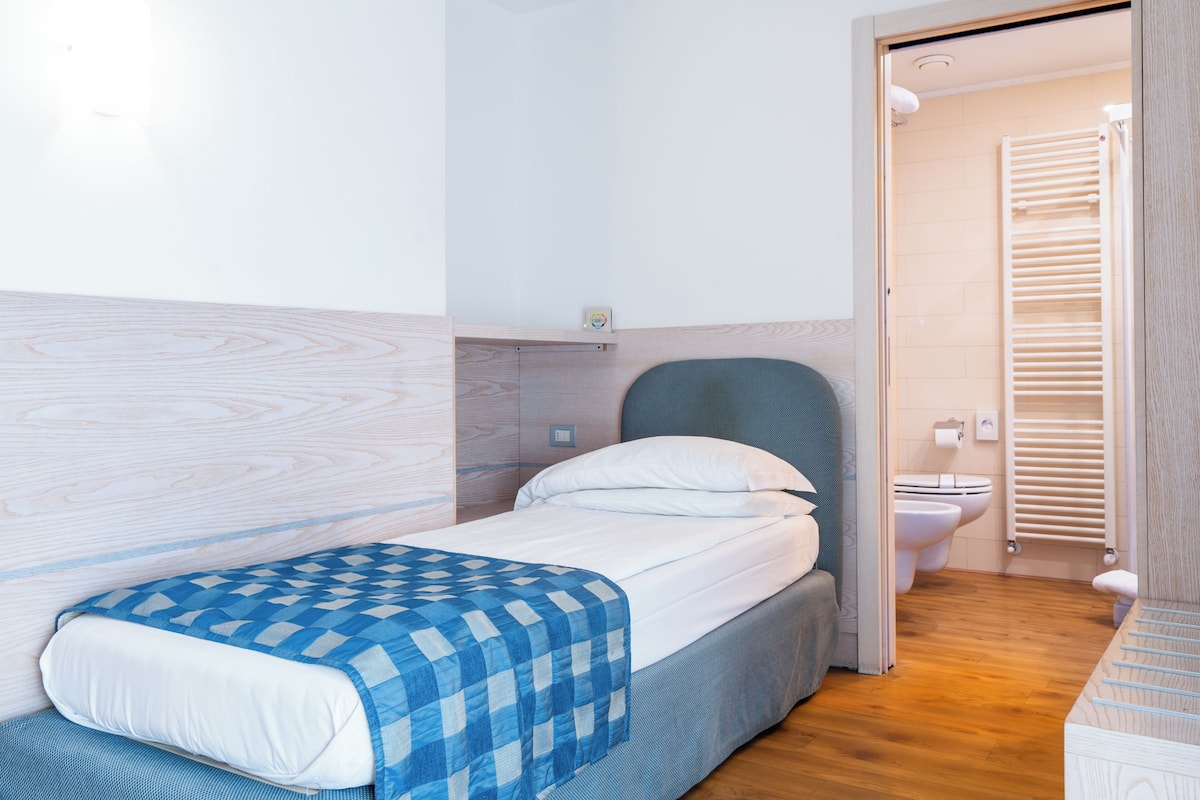 A bedroom with a bed, a bedside table, a wall-mounted shelf, and a view into the bathroom.