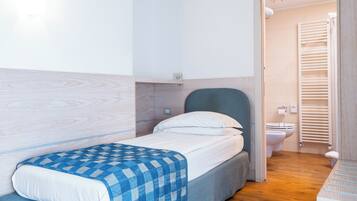 A bedroom with a bed, a bedside table, a wall-mounted shelf, and a view into the bathroom.