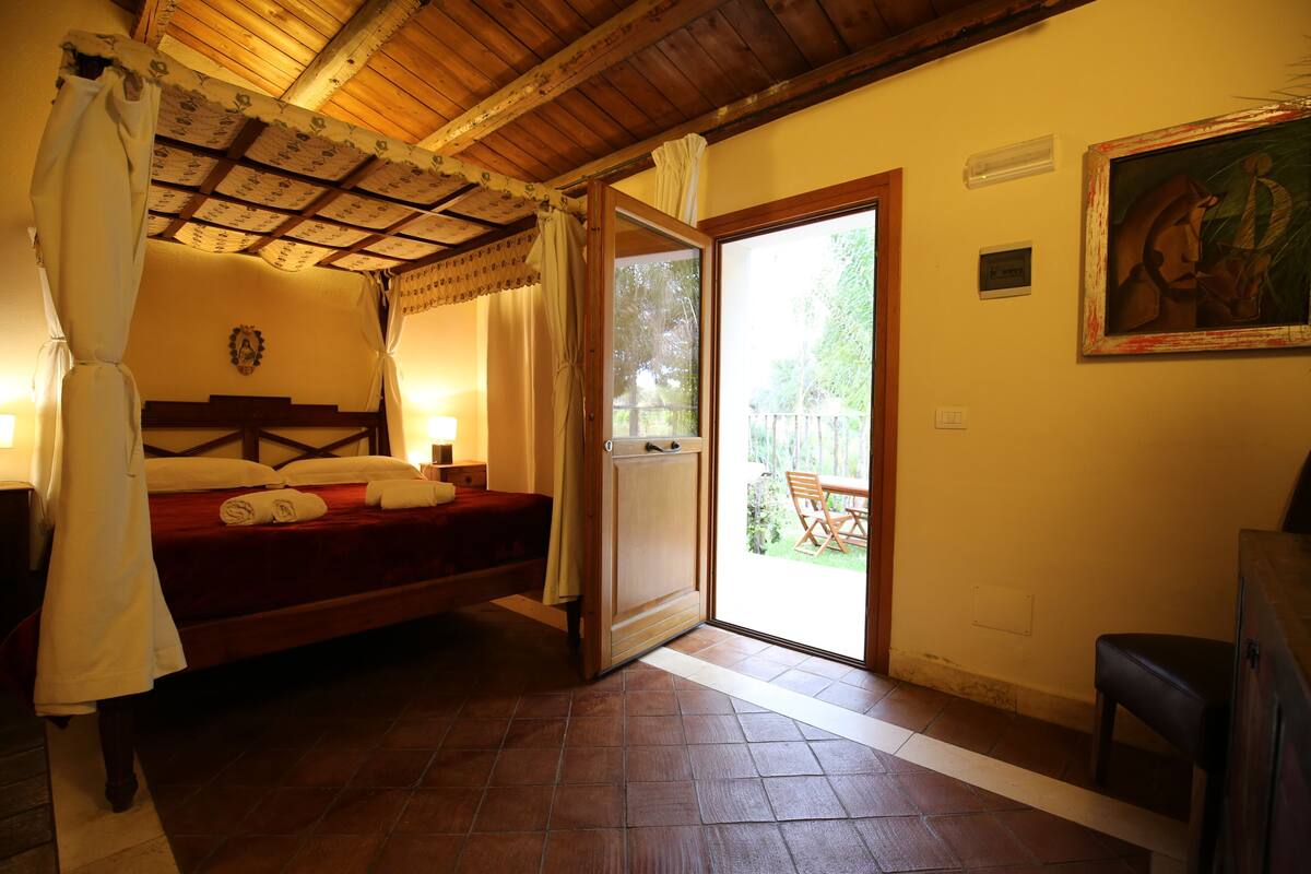 Un dormitorio con techo de madera, una cama con colcha roja, un dosel blanco y una puerta que da a un espacio al aire libre con una mesa y sillas.