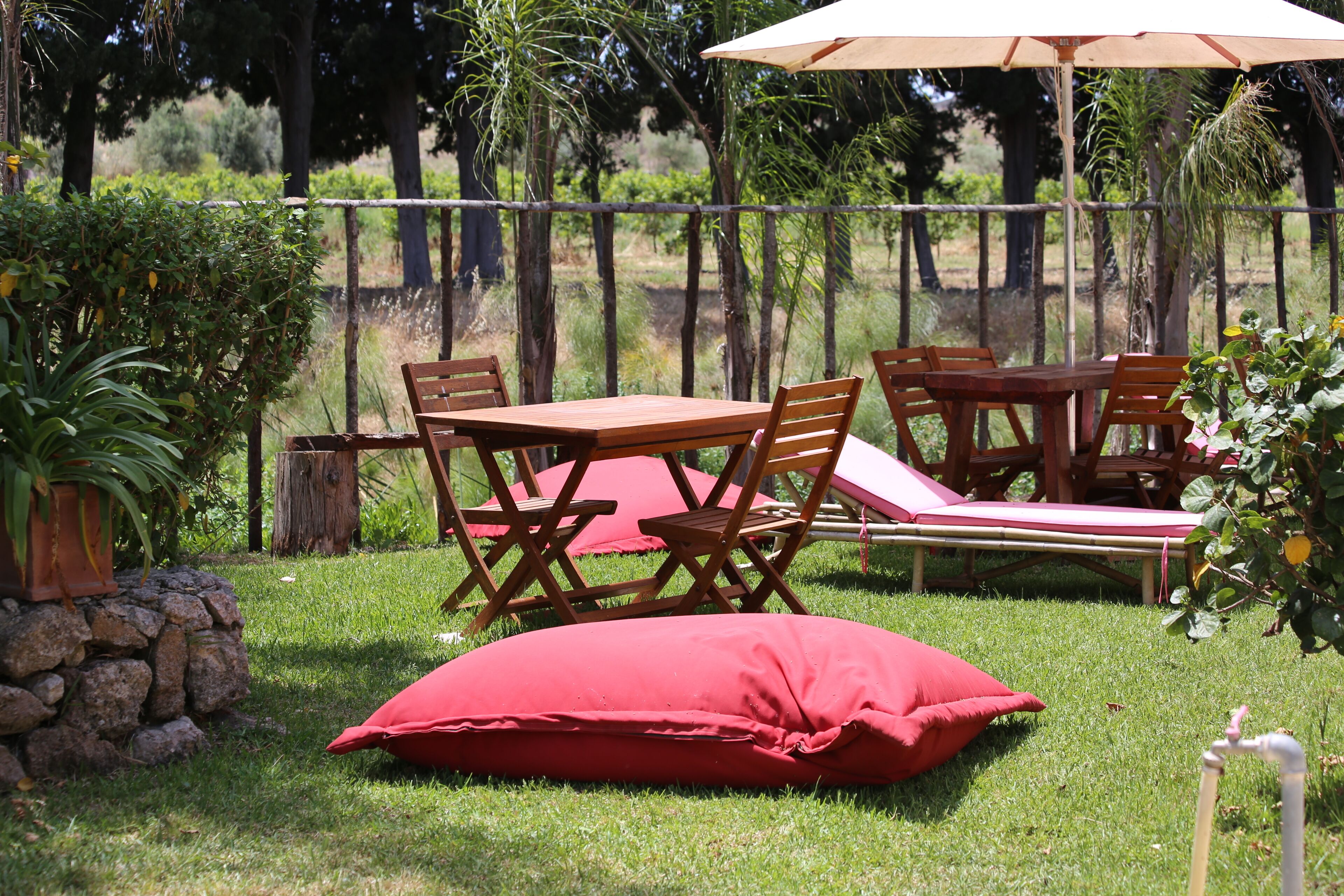Een terras buiten met houten meubels, waaronder een tafel, stoelen, een rode kussen en een houten kruk.