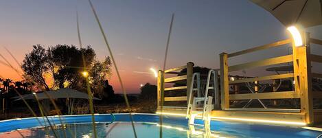 A swimming pool with a wooden deck and a ladder, surrounded by trees and evening sky.