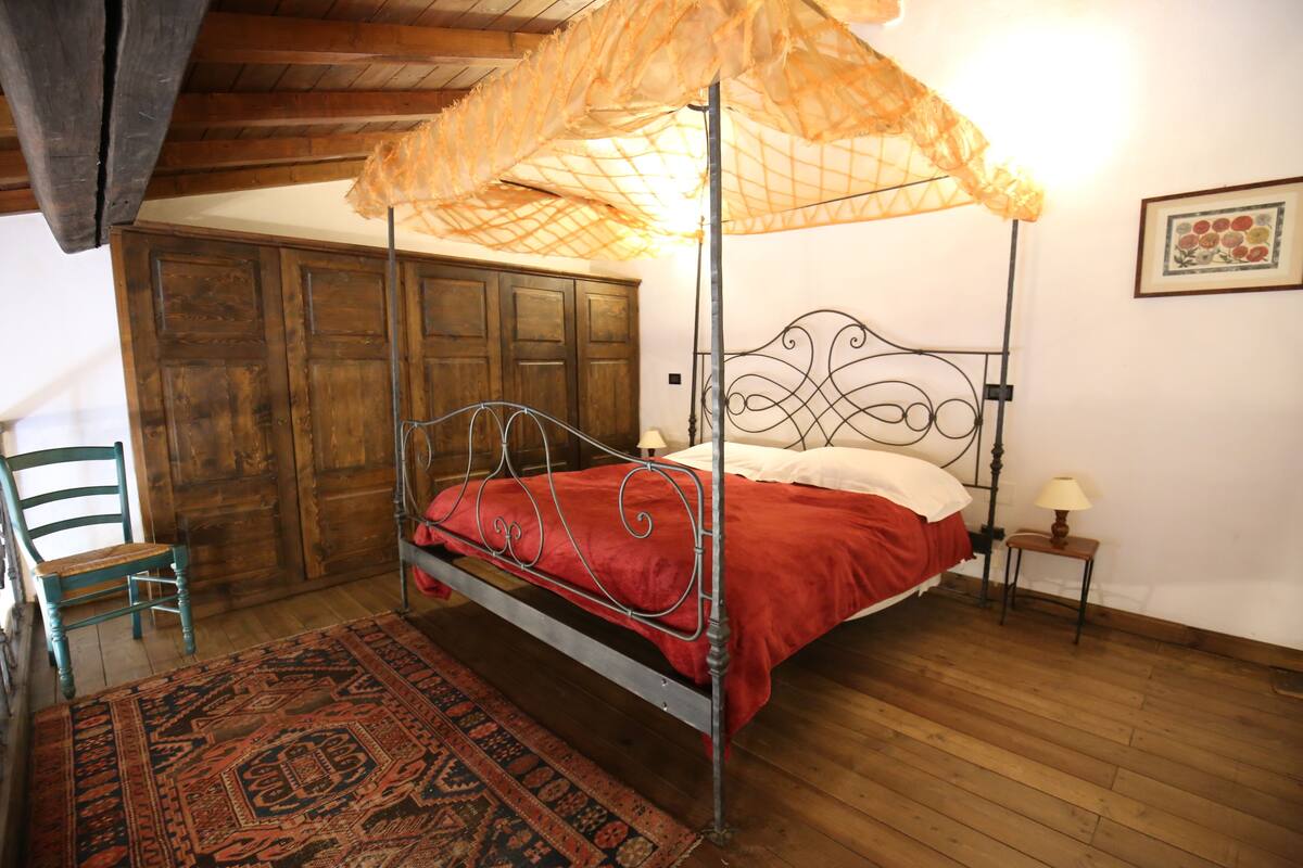 Una cama con dosel con colcha roja, suelo de madera, una silla y un armario de madera.