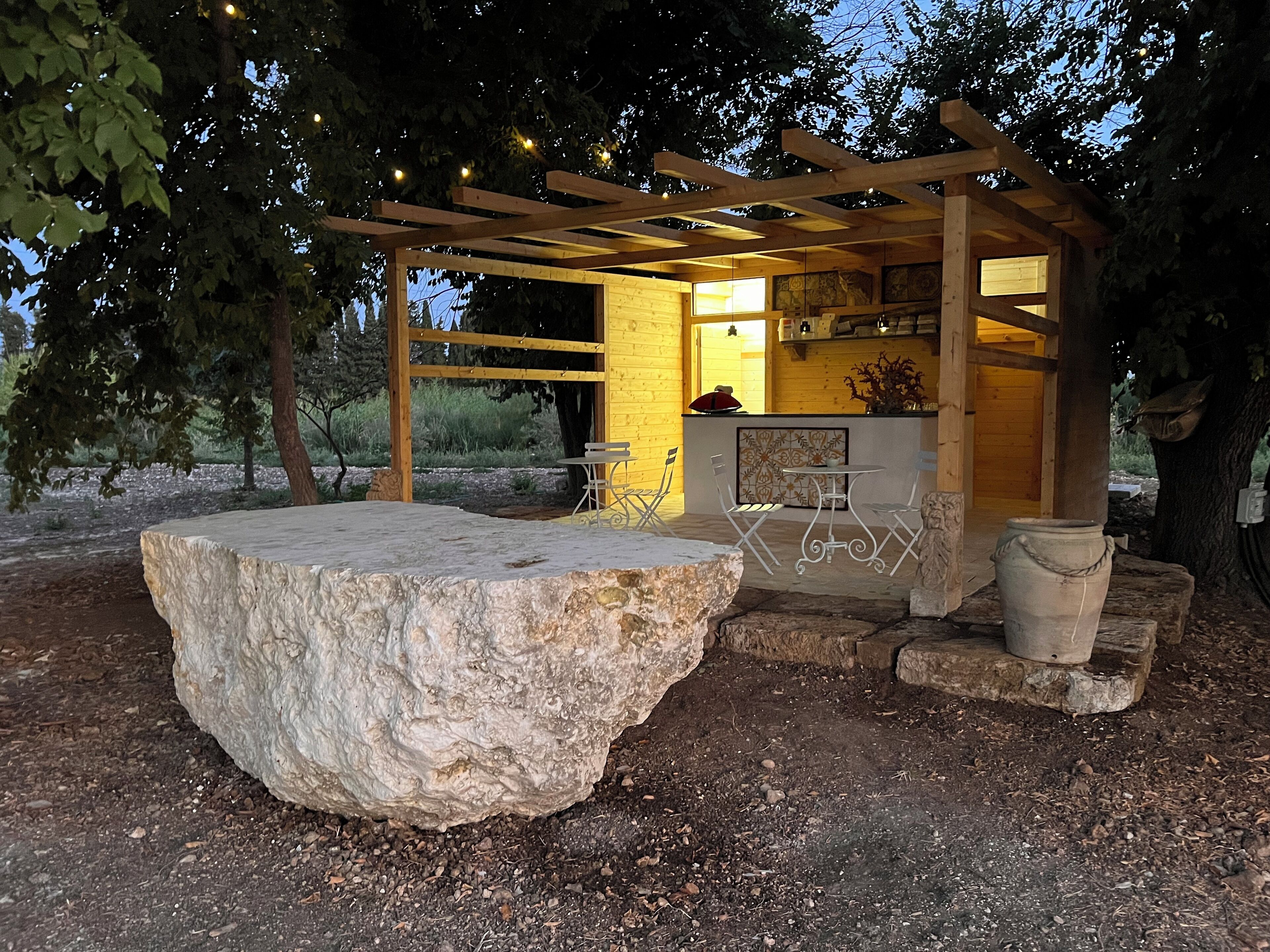 A rustic outdoor seating area with a large stone table, wooden pergola, and a small bar area with a counter and chairs.