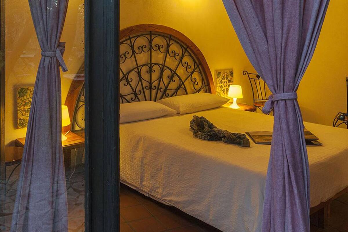 Un dormitorio con cama, cortinas, lámpara y cabecera decorativa.