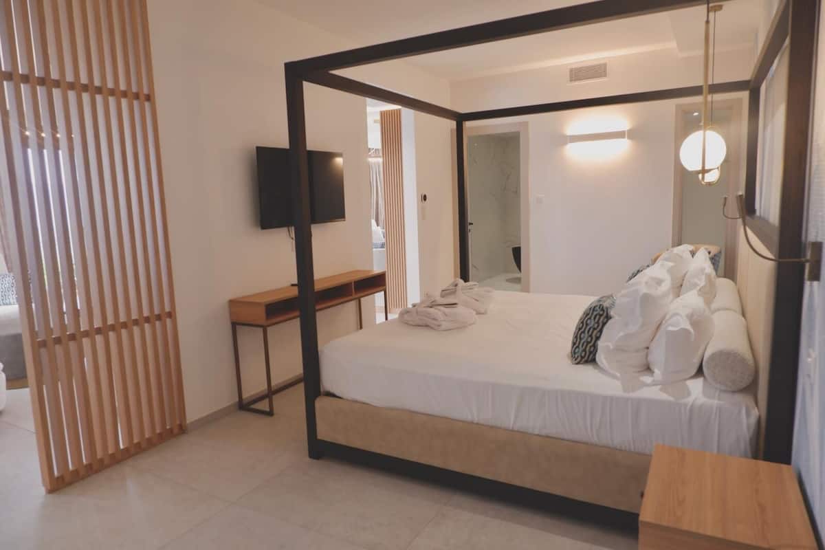 A modern hotel room with a four-poster bed, a flat-screen TV mounted on the wall, and a wooden desk.
