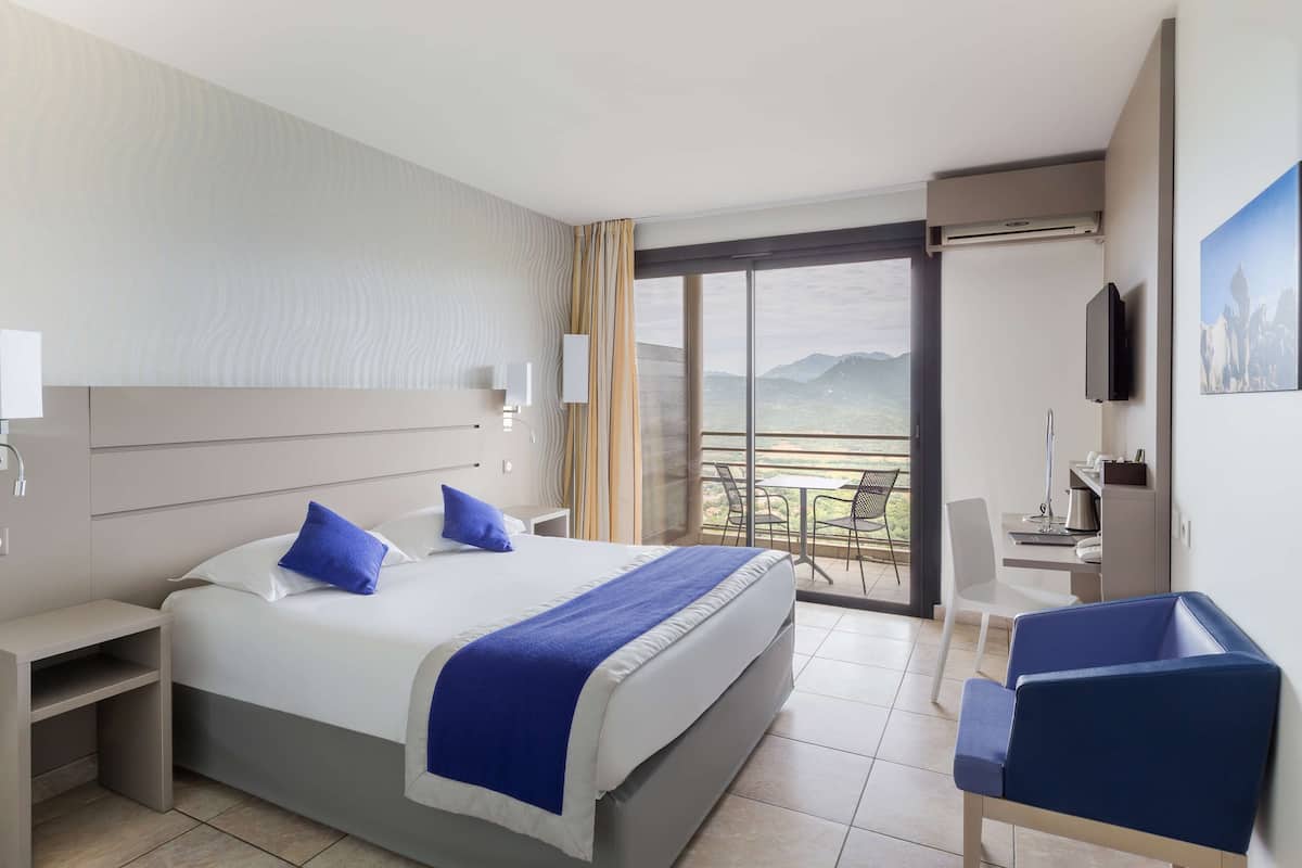 A hotel room with a large bed, a desk, a chair, and a balcony with a view.