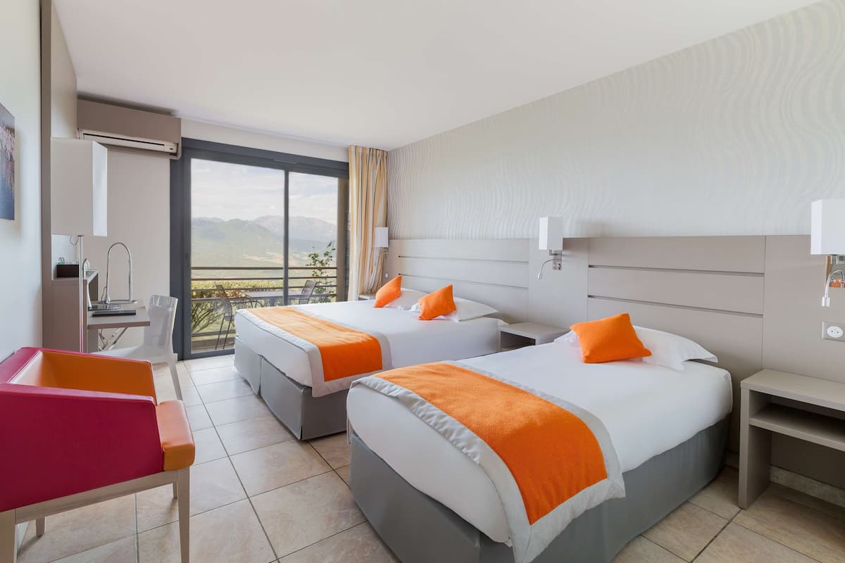 A hotel room with two beds, a desk, a chair, and a balcony with a view of the mountains.