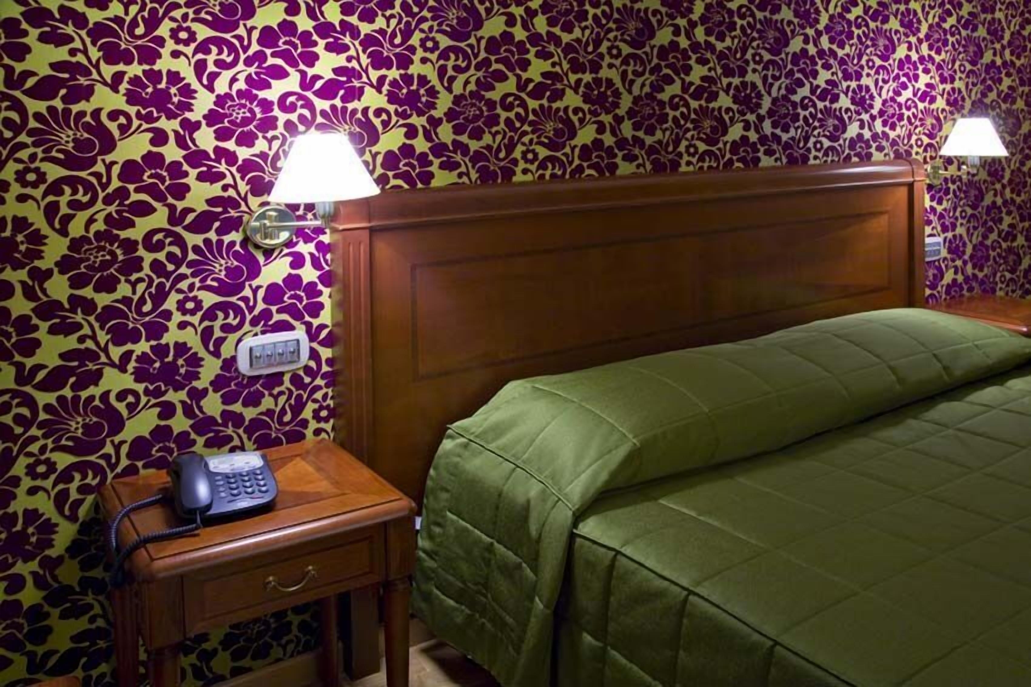 A hotel room with a floral-patterned wall, a wooden headboard, a green bedspread, a bedside table with a lamp, and a telephone.