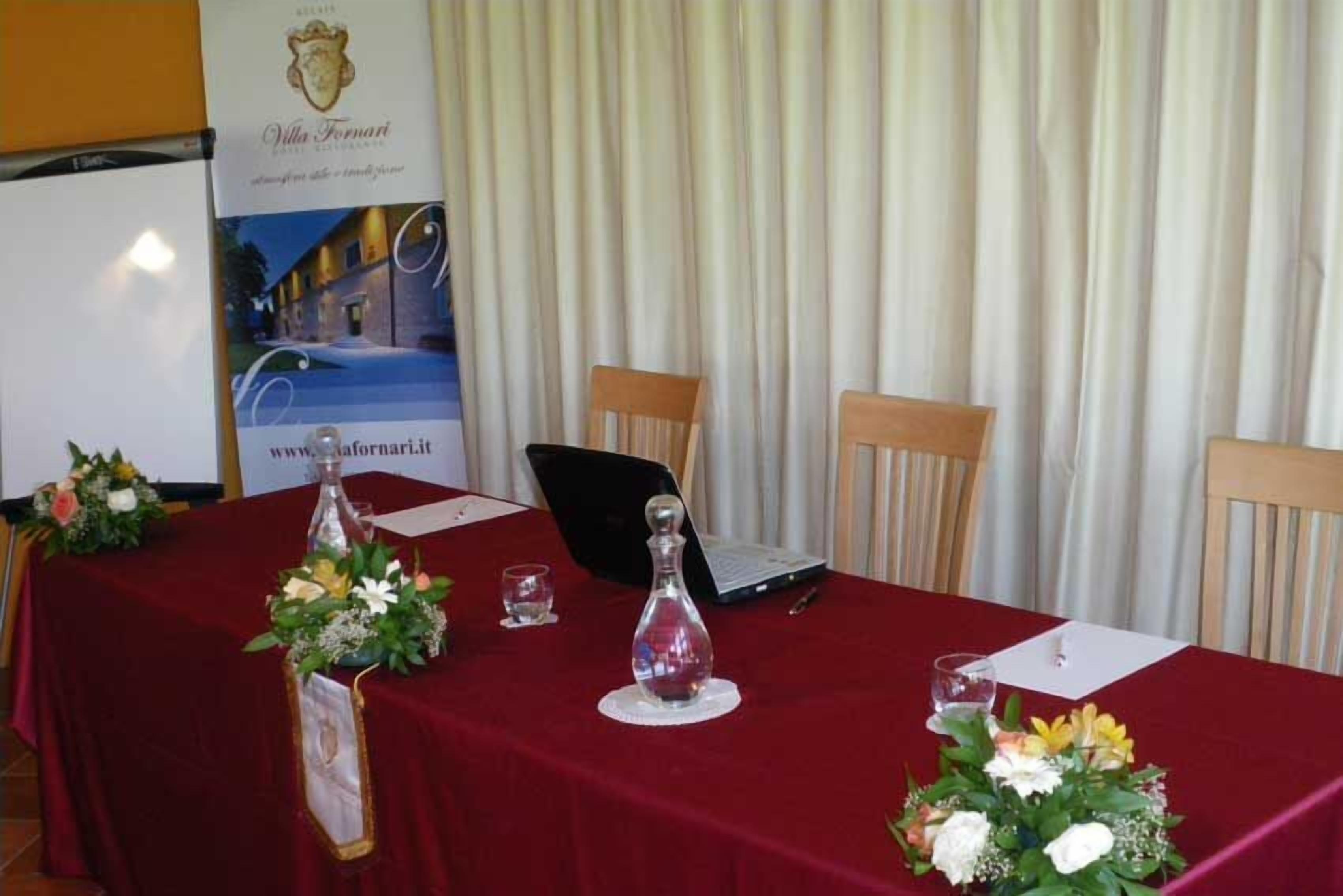 Una mesa de conferencias con mantel rojo, dos sillas, una computadora portátil, un decantador de vidrio y centros de mesa florales.