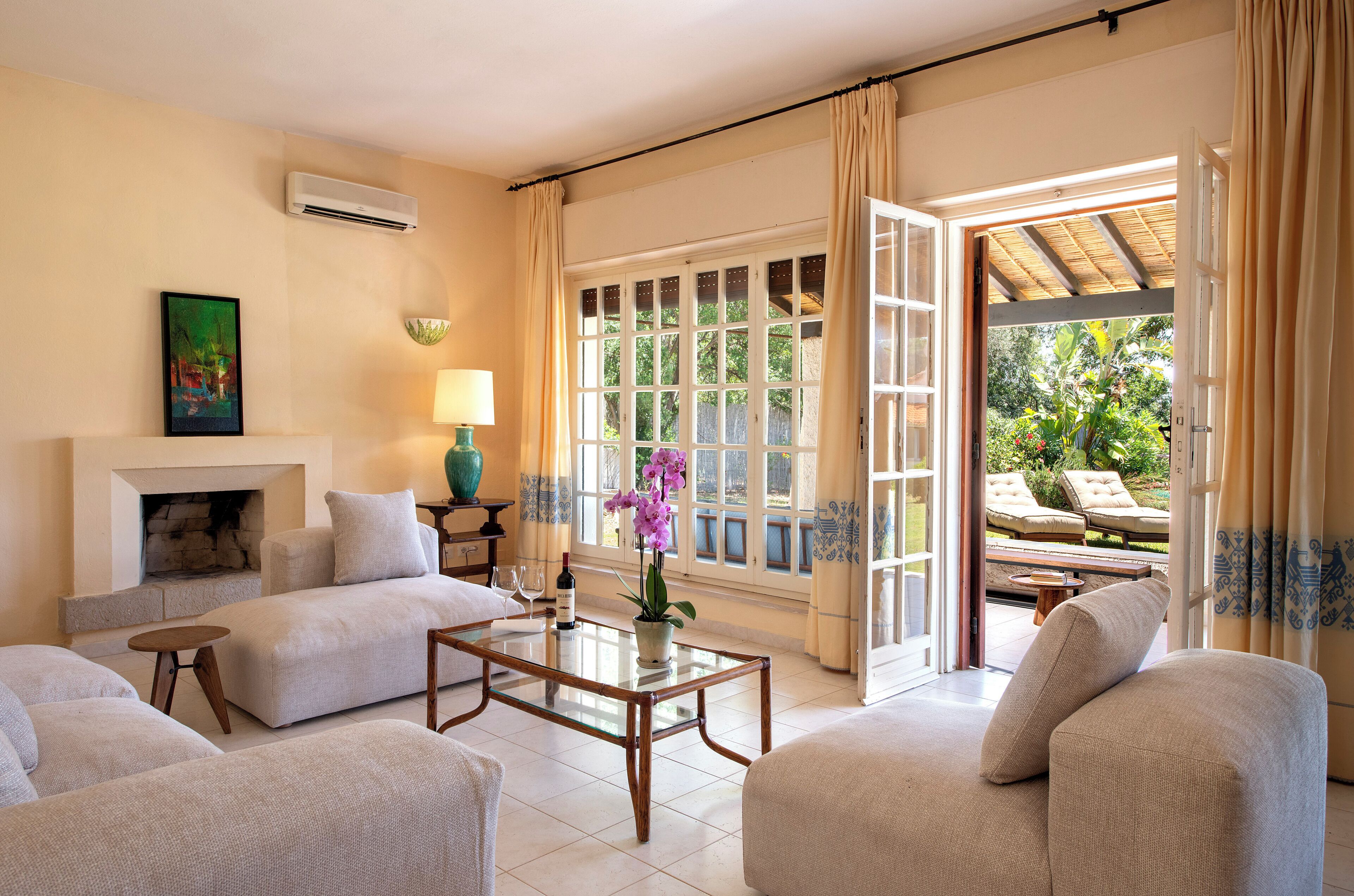 A living room with a fireplace, a glass coffee table, and a view of the garden through large windows.