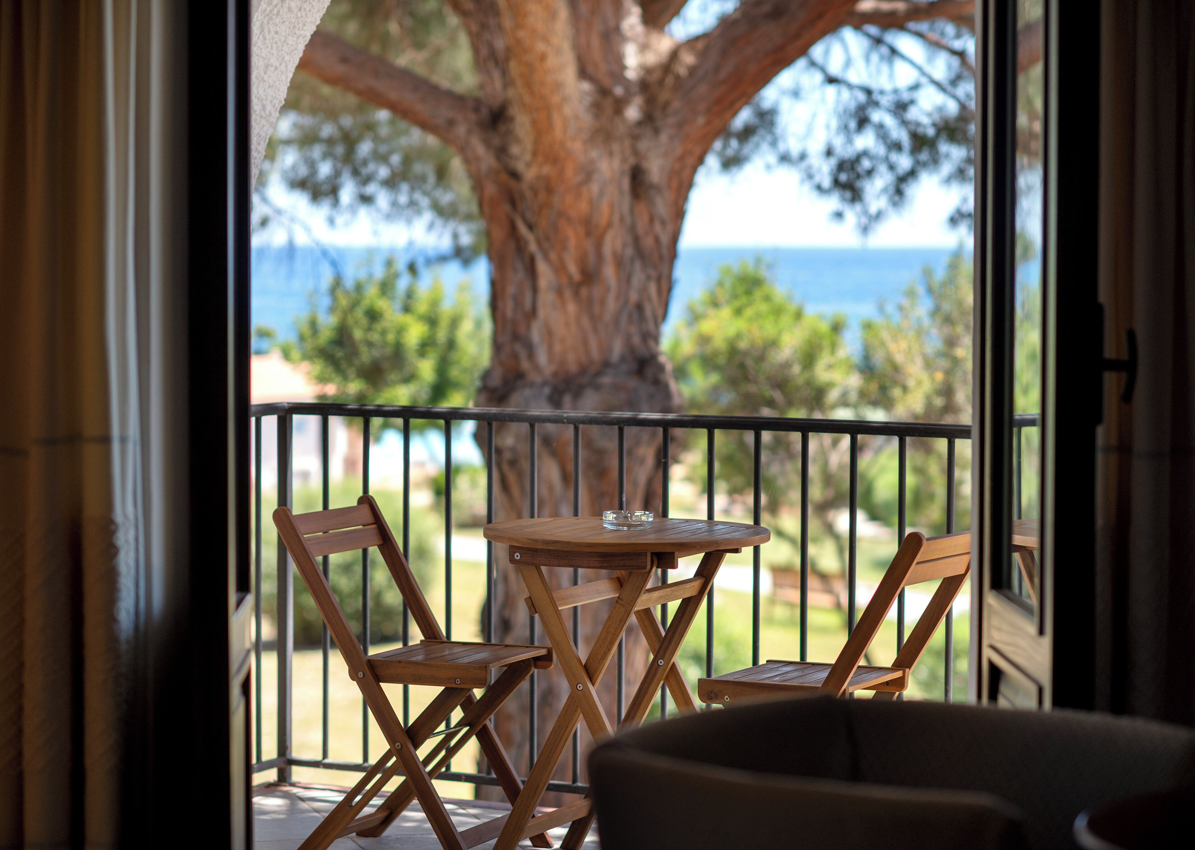 Un balcone con tavolo in legno e due sedie, affacciato su un albero secolare e vista mare.