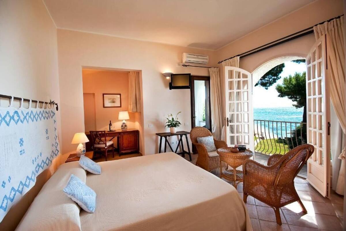 Habitación de hotel con cama, escritorio, silla, una mesita y vista al mar a través de una puerta abierta.