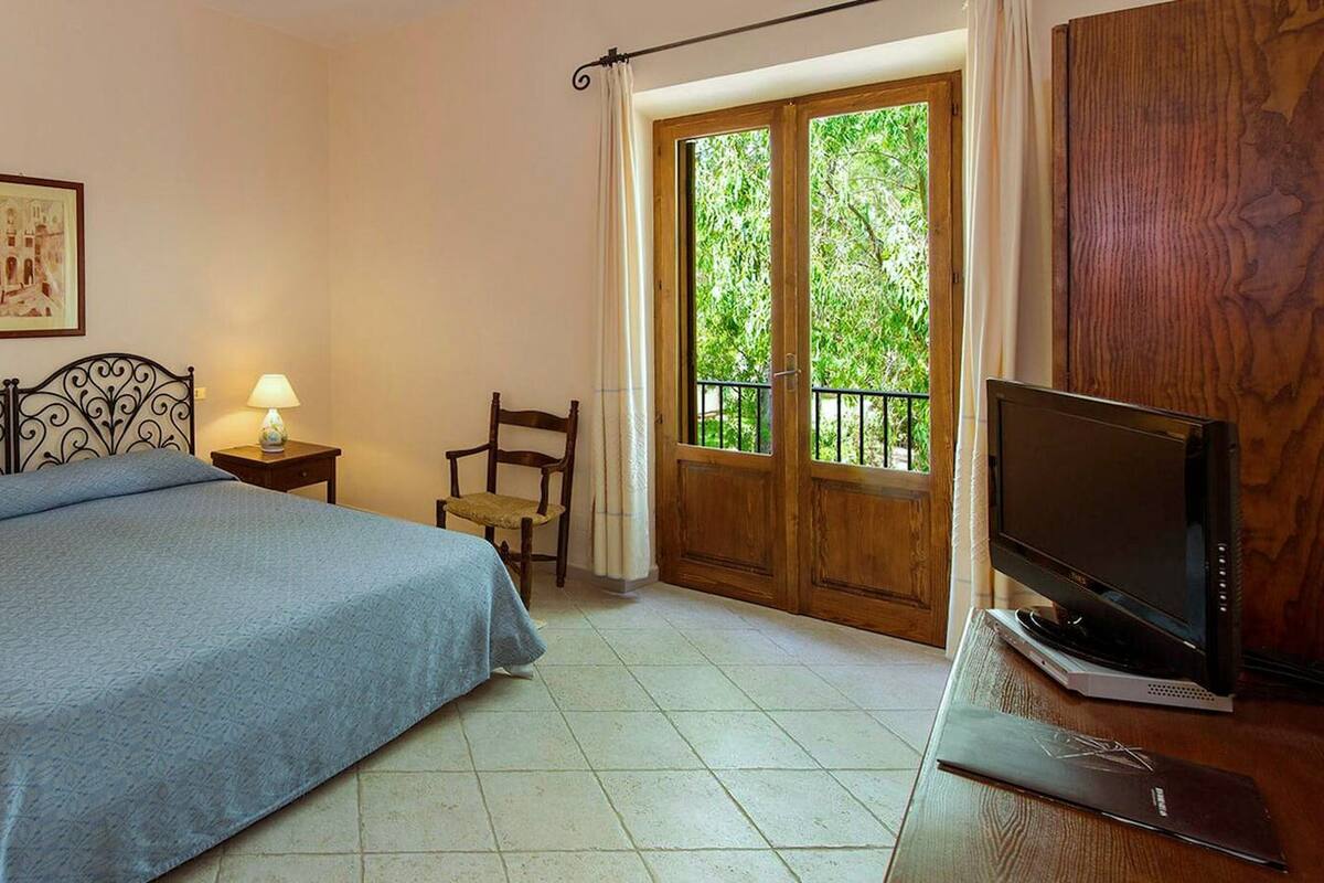 Un dormitorio con cama, mesitas de noche, una silla, un televisor y ventana con vistas a zonas verdes.