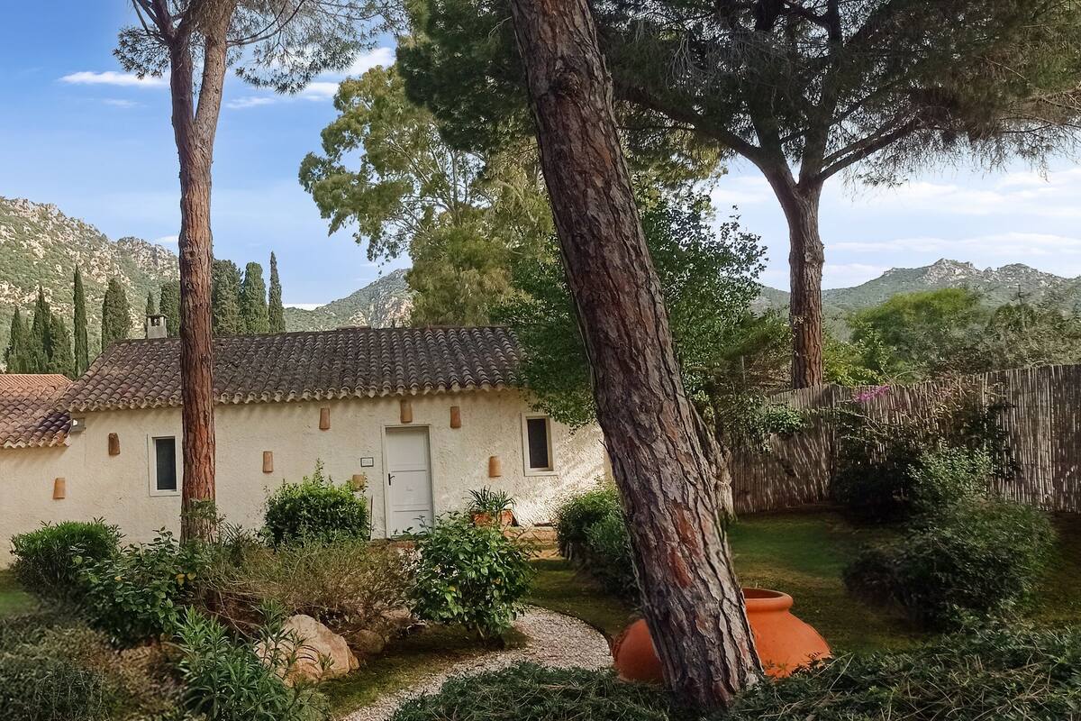 Una casa rústica con techo de tejas, rodeada de exuberante vegetación y montañas al fondo.
