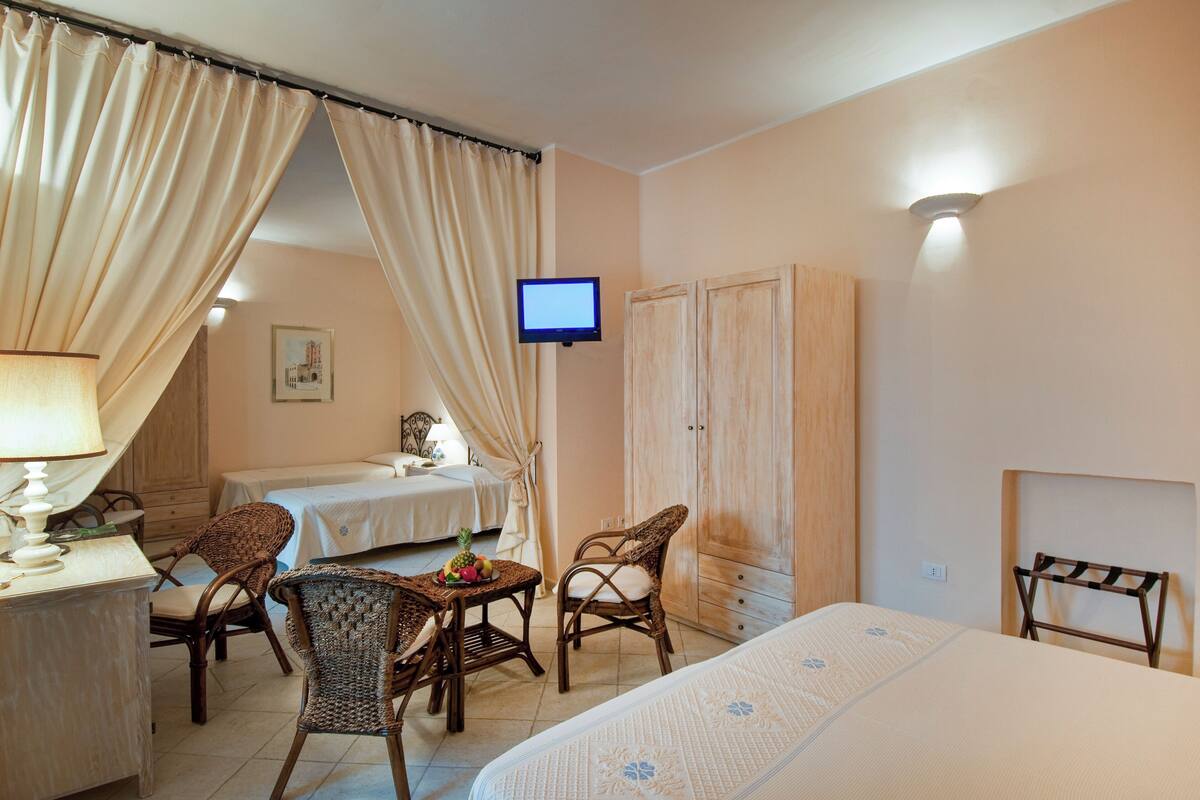 Habitación de hotel con cama, escritorio con lámpara, dos sillones de mimbre, una mesita con fruta, televisor y armario.