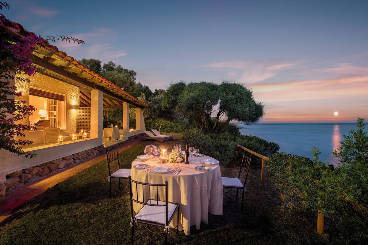 Un área de comedor al aire libre con una mesa preparada para cuatro personas, rodeada de sillas, cerca de un edificio con fachada de piedra y vista al mar al atardecer.
