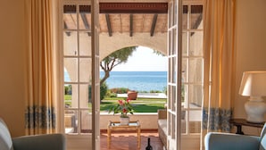 An open doorway leading to a patio with a view of the sea, a table with a bottle and glass, and two blue armchairs.