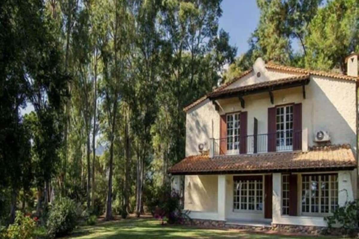 Una casa de dos pisos con techo de tejas, rodeada de un jardín exuberante y árboles altos.