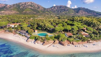 Une vue aérienne d’une station balnéaire, avec une mer d’un bleu limpide, une plage de sable et une piscine entourée d’une végétation luxuriante, le tout sur fond de montagnes.