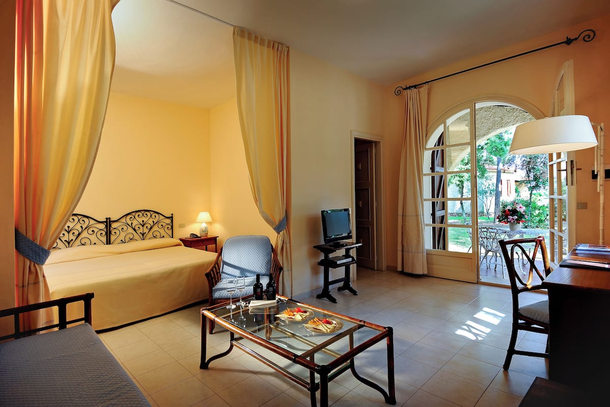 Habitación de hotel con cama, sofá, televisión, mesa de comedor con comida y ventana con vistas.