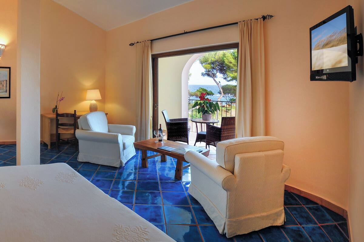 Habitación de hotel con cama, dos sillas, una mesa, una lámpara, un cuadro y vista al mar.