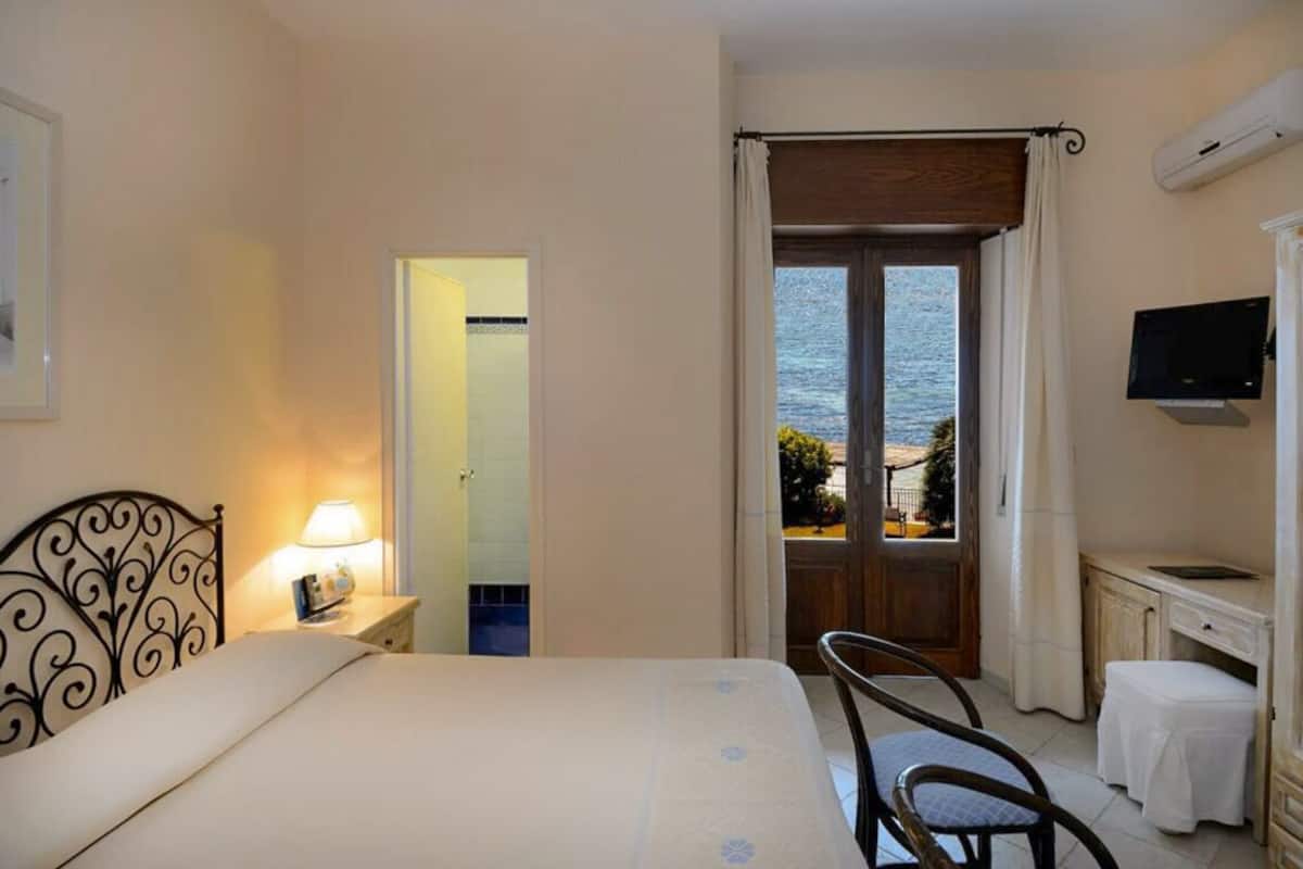 Habitación de hotel con cama, una silla, un escritorio, televisor y vista al mar a través de una puerta.