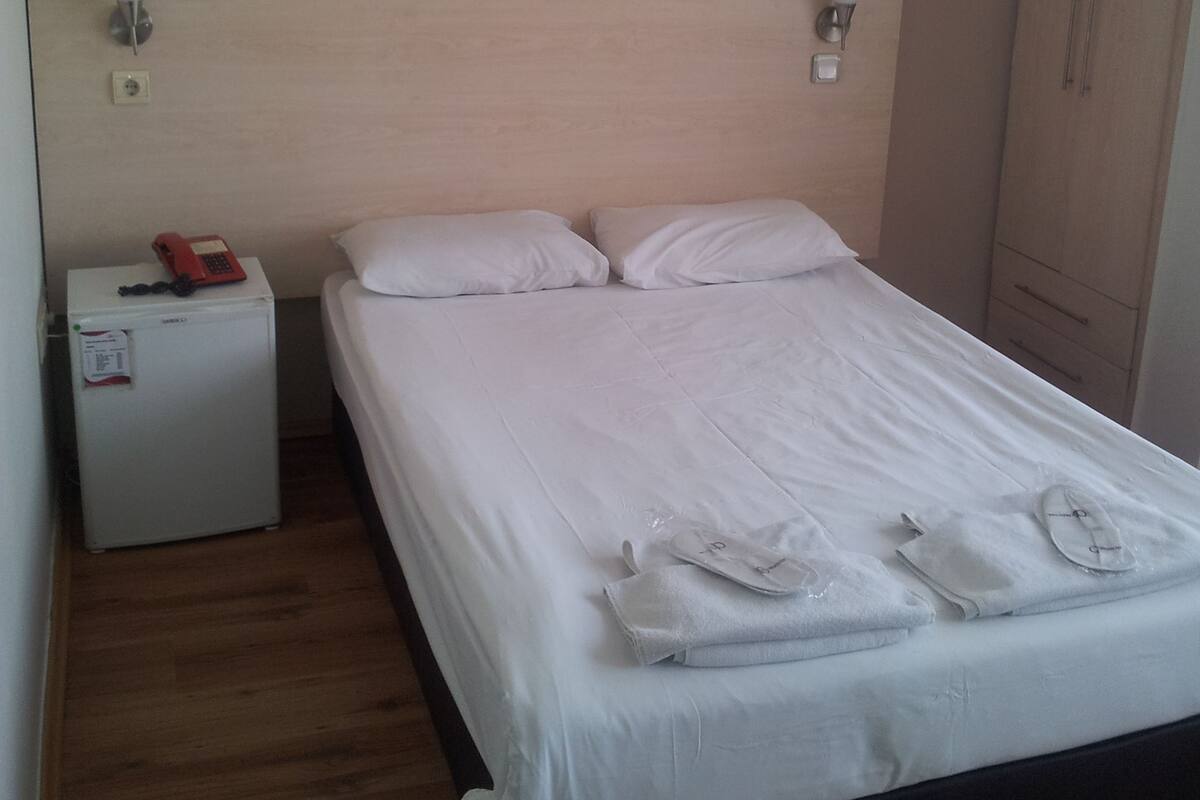 A neatly made bed with white linens and pillows, a small bedside table with a red telephone, and a wall-mounted light fixture.