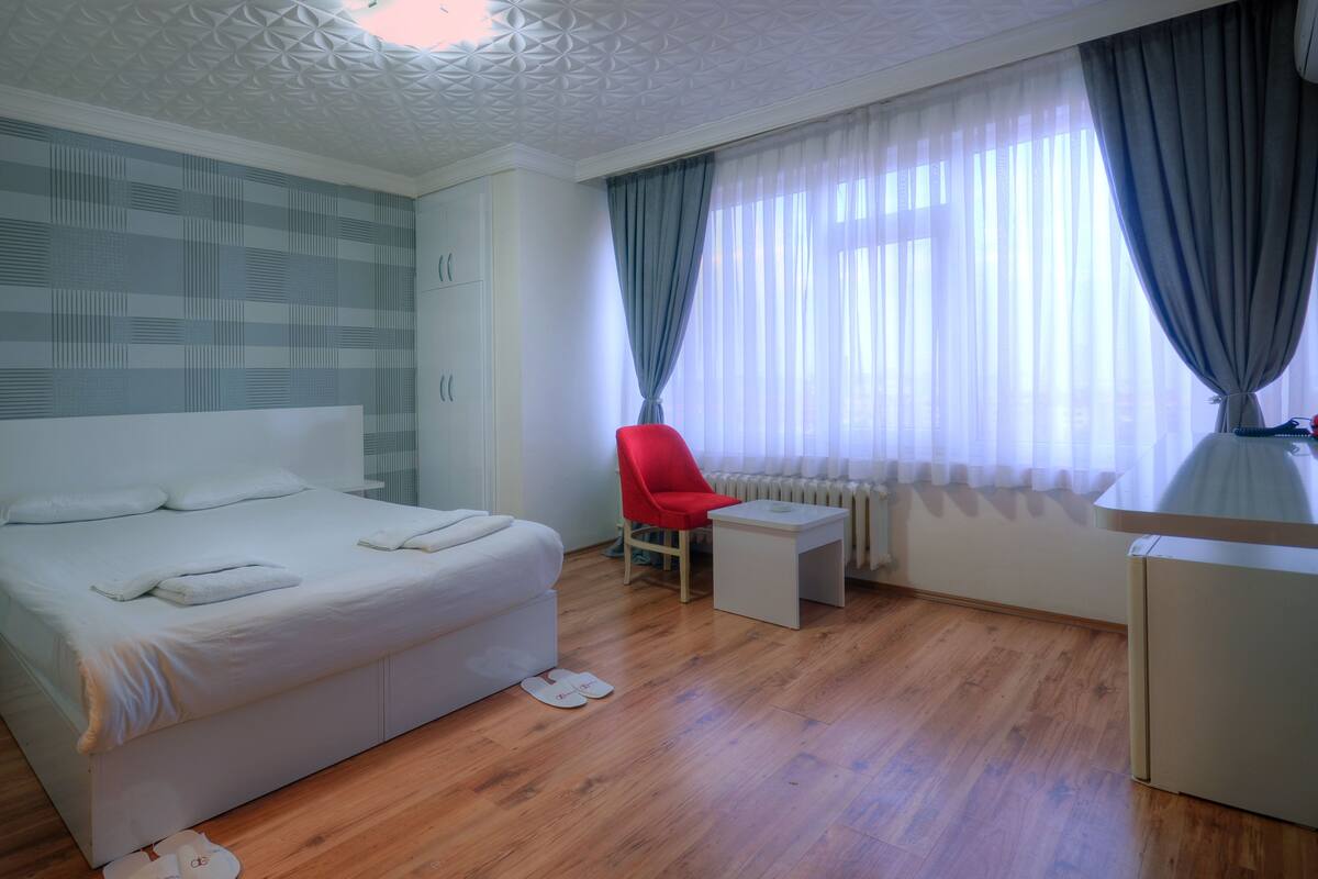 A modern hotel room with a large bed, a small desk, a red chair, and a window with sheer curtains.