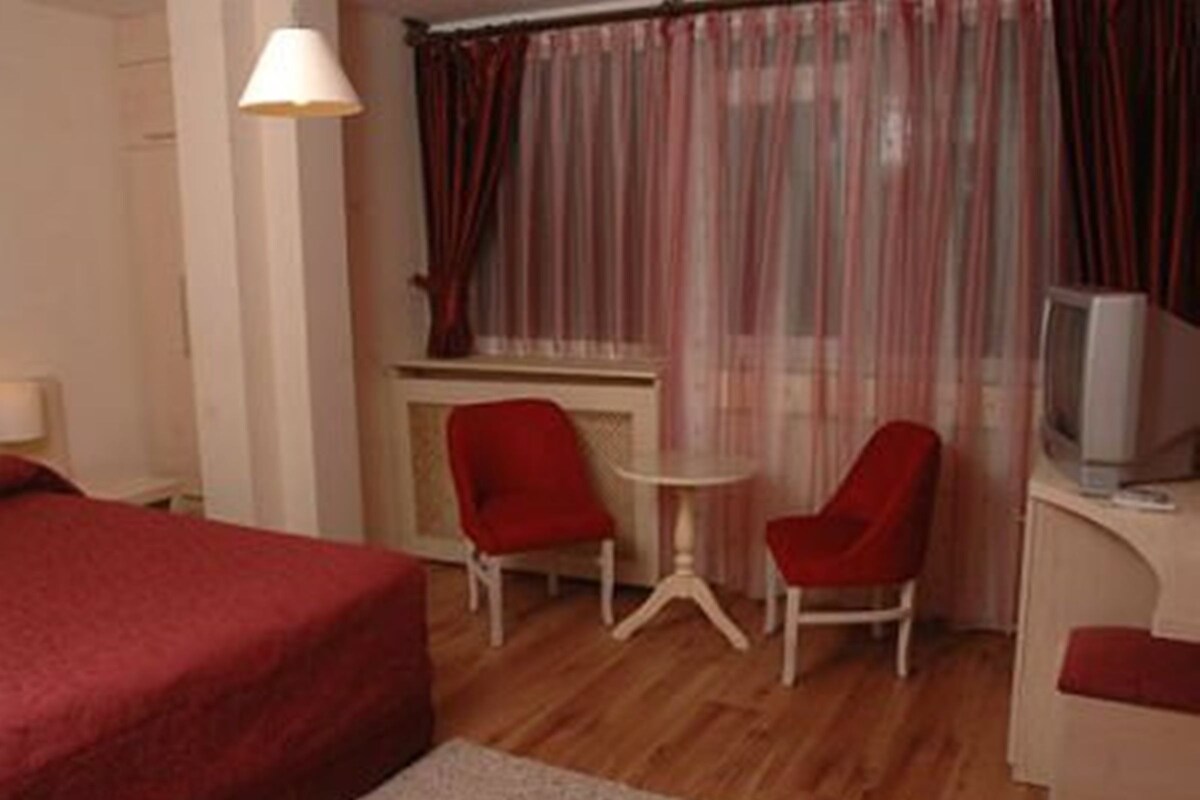 A hotel room with a bed, two red chairs, a small table, a television, and a window with curtains.