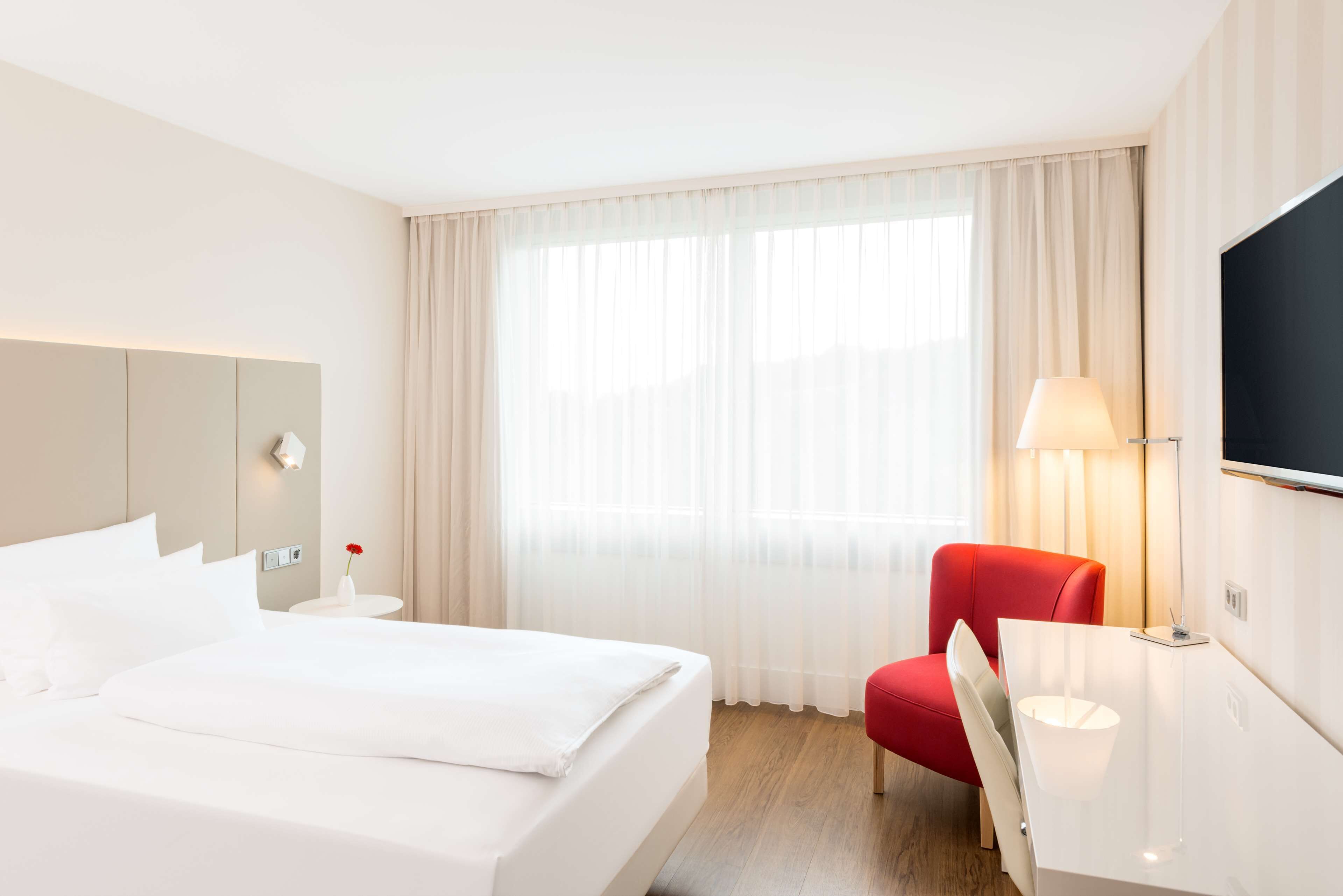 A hotel room with a large bed, a red chair, a desk with a lamp, a TV on the wall, and a window with sheer curtains.