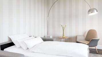Una moderna camera d'hotel con un letto rifatto, una poltrona grigia, un tavolino con un vaso di fiori e una lampada da terra ad arco.