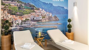 A hotel room with a large scenic wall mural, two lounge chairs, a small table with a drink, and a potted plant. - NH Collection Köln Mediapark (Cologne)