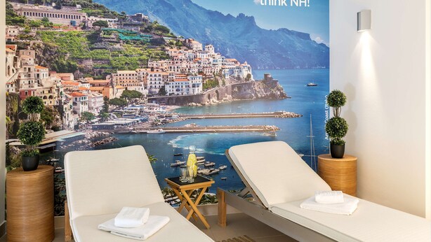 Habitación de hotel con un mural panorámico grande, dos sillones de descanso, una mesita con una bebida y una planta en maceta.