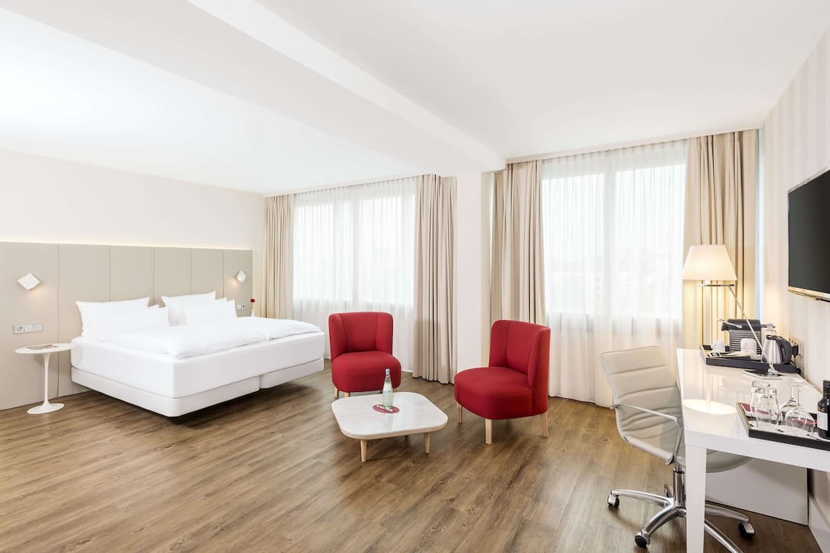 Ein Hotelzimmer mit einem Bett, zwei roten Sessel, einem Schreibtisch mit Lampe und einem Couchtisch.